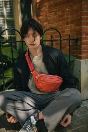 A stylish person sits outdoors with a red belt bag, gray pants, black jacket, and Adidas shoes, posing against a brick wall.