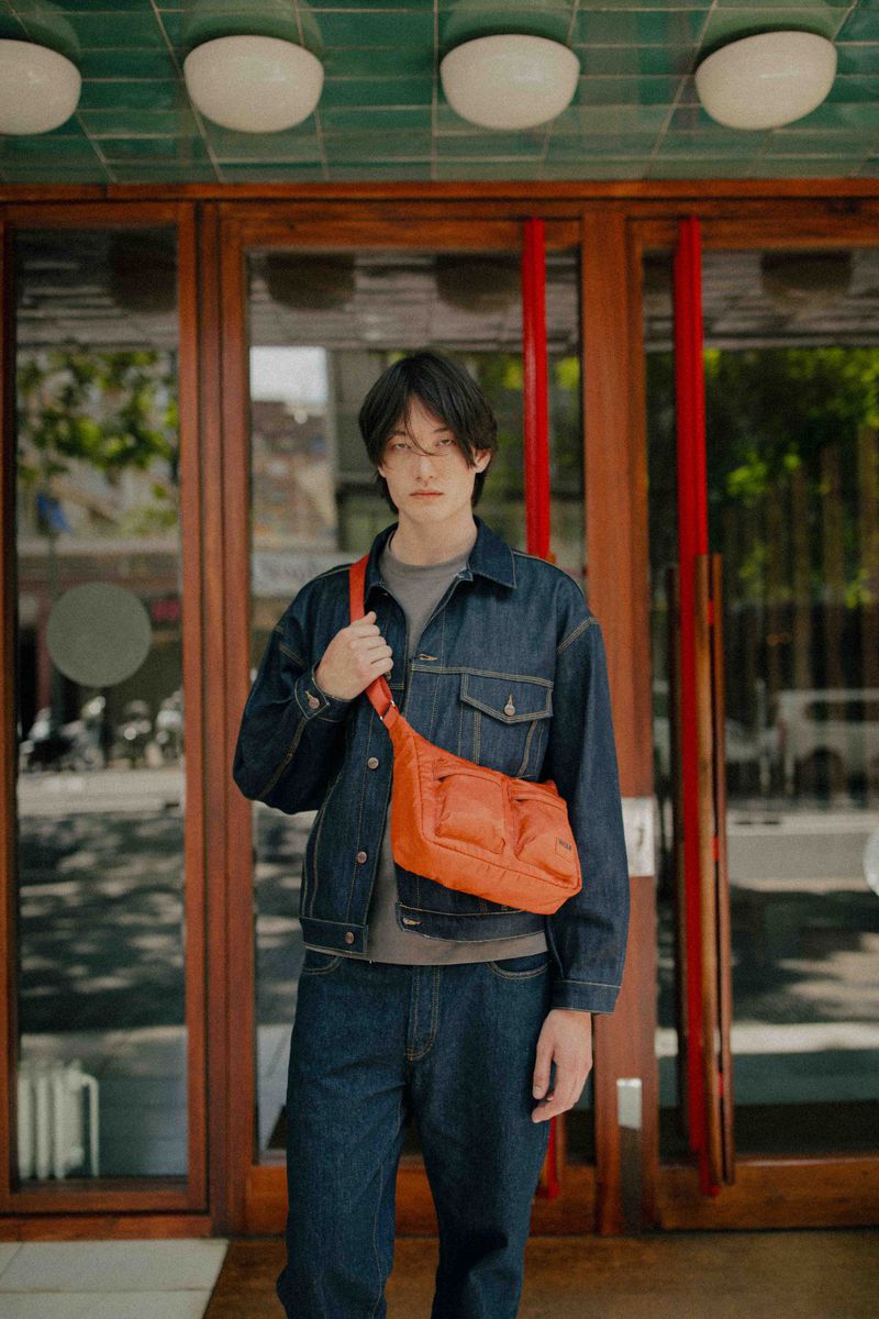 A stylish young adult in a denim jacket and jeans stands in front of a building wearing an orange crossbody bag, showcasing a casual fashion look.