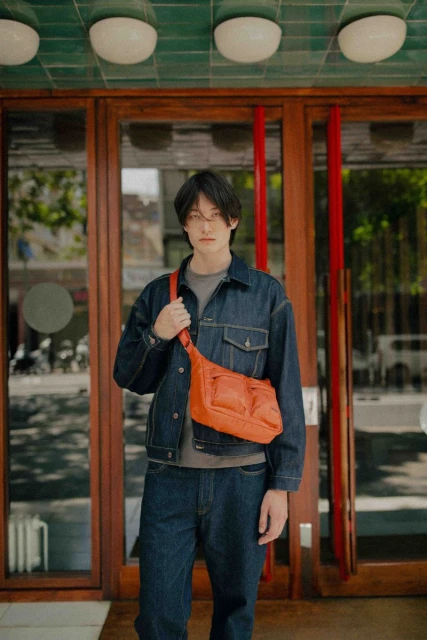 A stylish young adult in a denim jacket and jeans stands in front of a building wearing an orange crossbody bag, showcasing a casual fashion look.