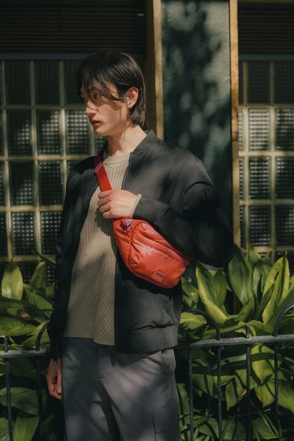 A young adult man poses outdoors, wearing a black jacket and grey trousers, with a bright red puffy shoulder bag in natural light.