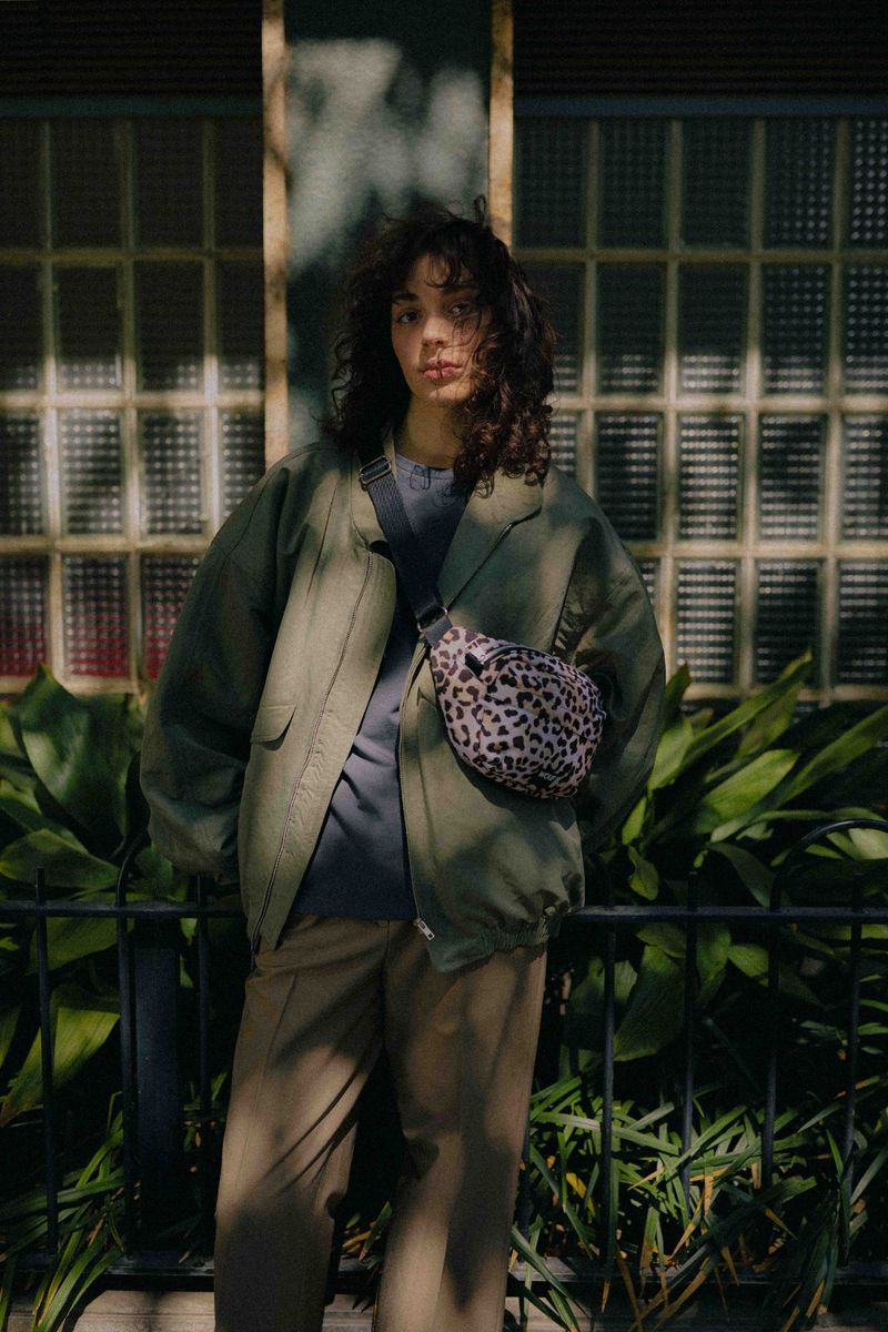 Adult woman with curly hair poses in olive green jacket and cargo pants with animal print bag in front of green foliage.