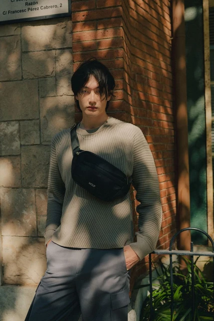 Portrait of a young Asian man in a neutral sweater with a black crossbody bag, posing against a brick wall in natural sunlight.