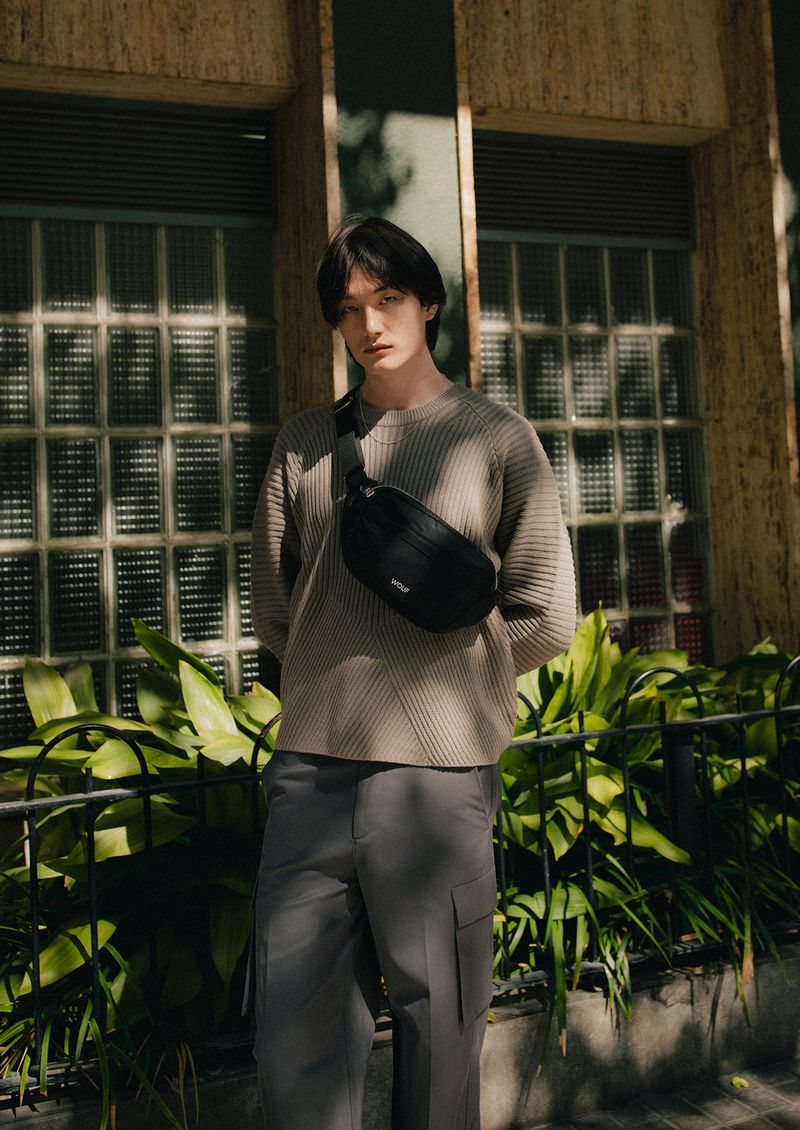 A solitary Asian male model with dark hair is the focal point in an outdoor setting, wearing a neutral-toned ribbed sweater and gray cargo pants.