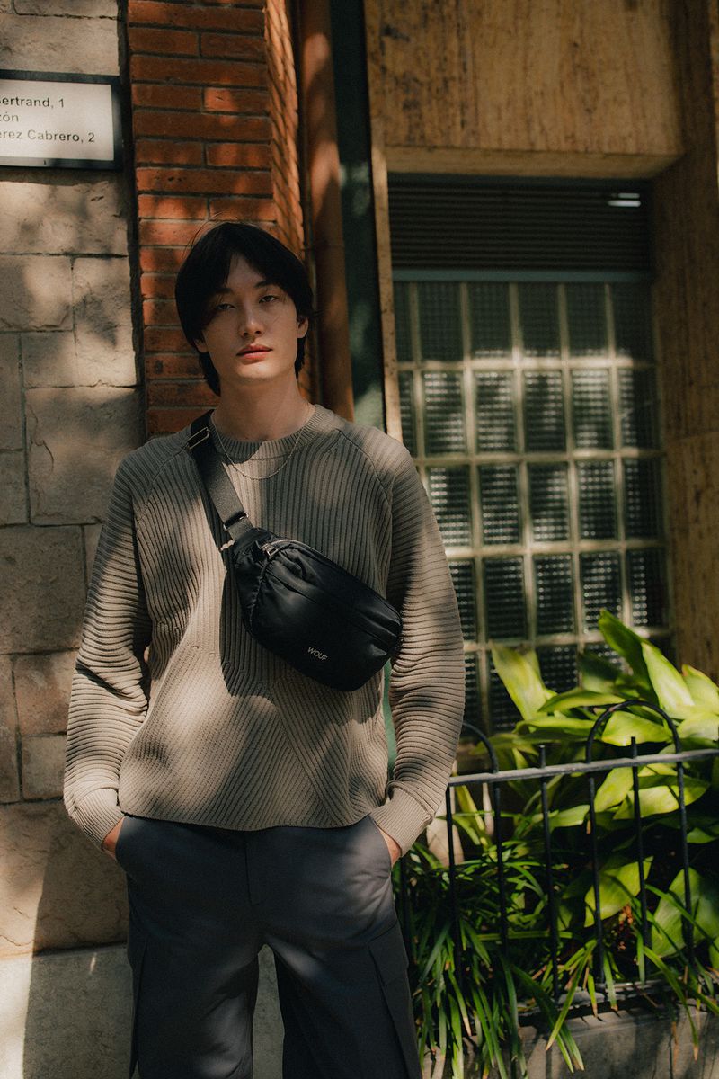 A young man wearing a grey ribbed sweater and dark trousers posing with a black crossbody bag outdoors in front of a brick building.