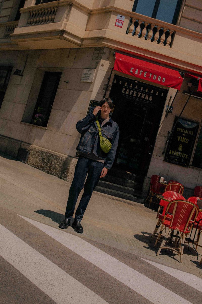 Adult man stands on crosswalk in front of cafe, wearing dark denim jacket, pants, black shoes, and green shoulder bag, looking at his phone.