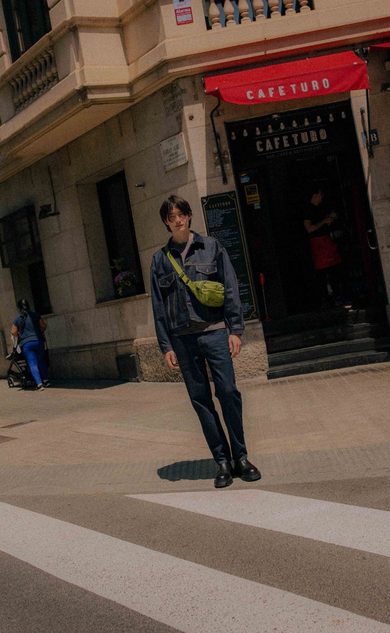 A young adult man in a denim jacket and crossbody bag stands on a crosswalk in front of a building with a red awning.