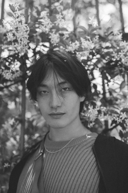 Portrait of a young adult with dark hair looking directly at the camera with a white-flowered bush in the background, black and white.