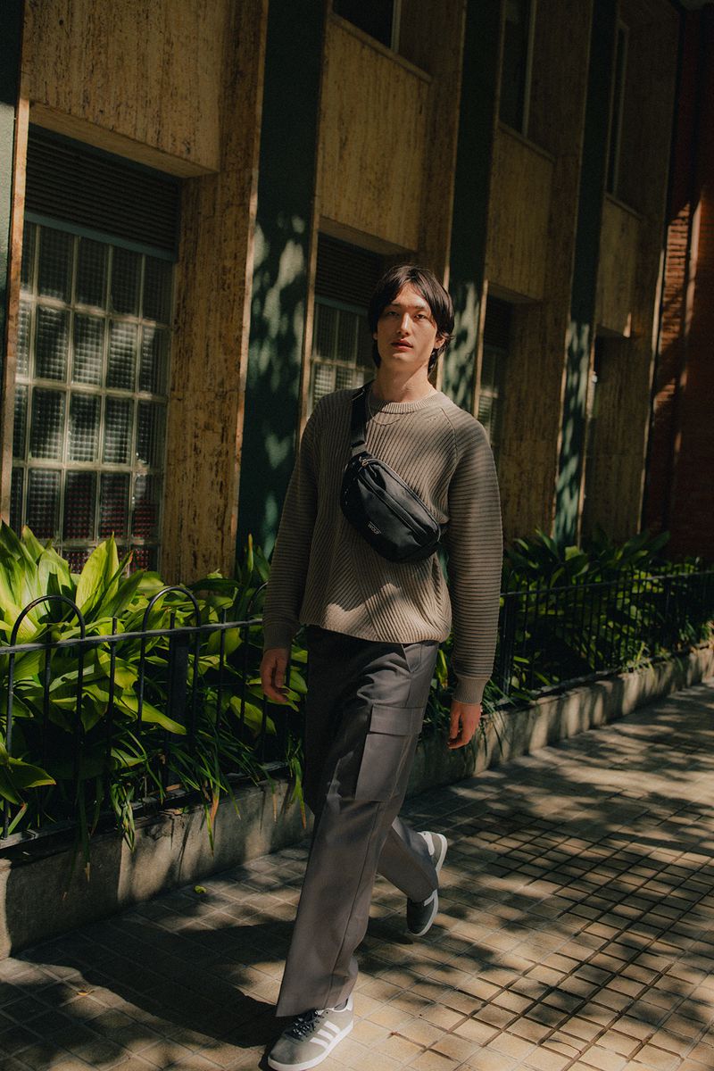Full-length shot of a young adult man walking on a city sidewalk wearing casual gray sweater, pants, and sneakers, and a black fanny pack.