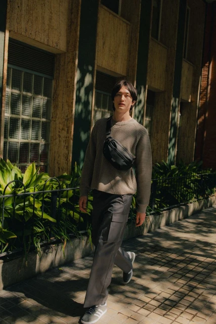 Full-length shot of a young adult man walking on a city sidewalk wearing casual gray sweater, pants, and sneakers, and a black fanny pack.
