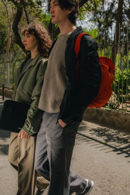 Two people walk on a sunny street. A woman carries a laptop and wears a green jacket; a man has a red backpack and a black jacket.
