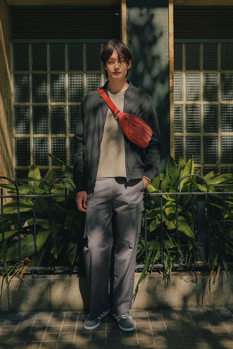 Stylish Asian man stands outdoors in a jacket, cream sweater, grey trousers, and grey sneakers, carrying a red fanny pack across his body.