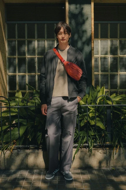 Stylish Asian man stands outdoors in a jacket, cream sweater, grey trousers, and grey sneakers, carrying a red fanny pack across his body.