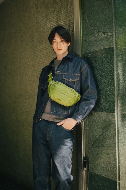 A stylish Asian male model leans against a textured wall in a denim jacket with a light green crossbody bag for a fashion editorial.