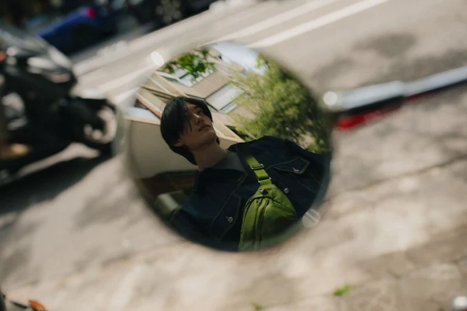 Reflection of a young adult male in a motorcycle mirror, wearing a denim jacket and crossbody bag on a sunny city street.
