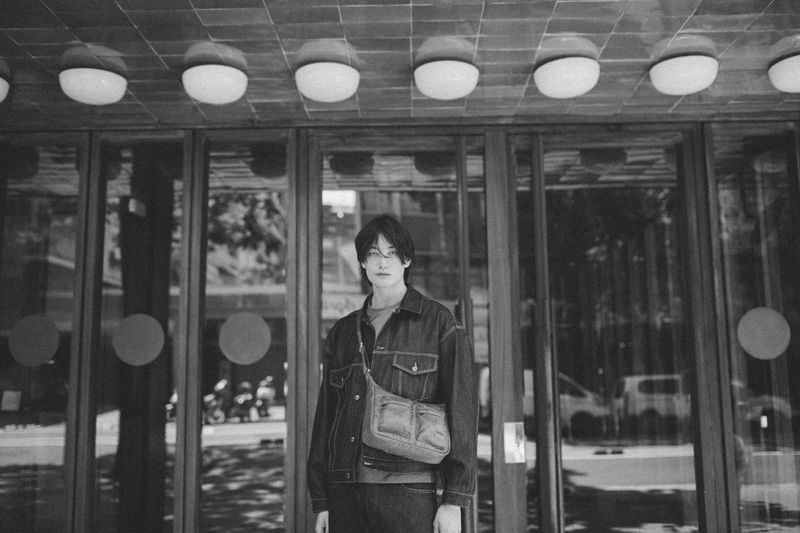 Monochrome portrait of a young adult with a denim jacket and shoulder bag standing in front of a glass-door building.