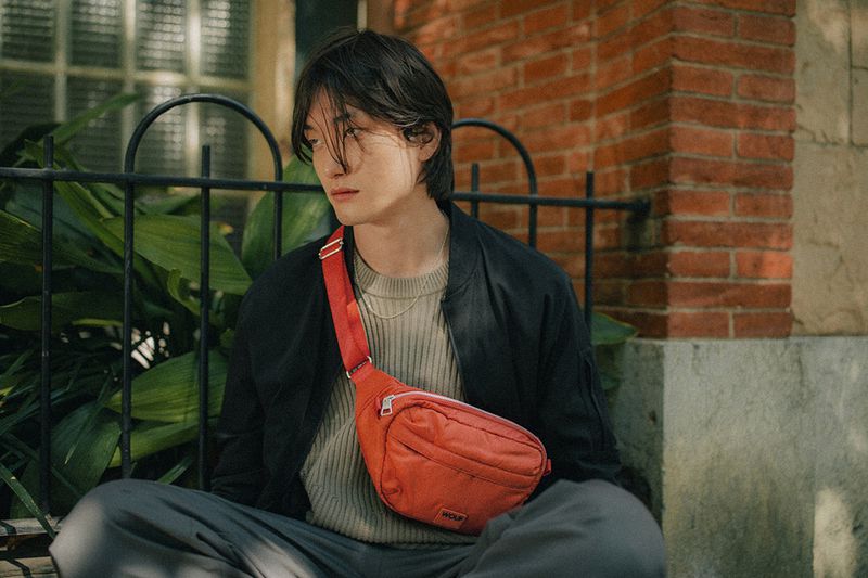 A person with dark hair sits outside on a bench, wearing a black jacket, gray pants, and an orange belt bag against a brick wall.