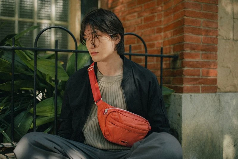 A person with dark hair sits outside on a bench, wearing a black jacket, gray pants, and an orange belt bag against a brick wall.