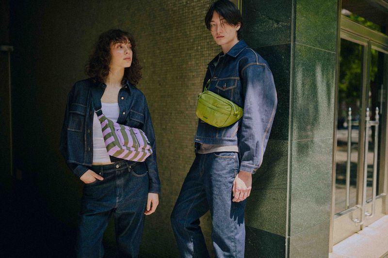 Two young adults, a man and woman, pose outdoors in matching denim outfits, each wearing a different color crossbody bag.