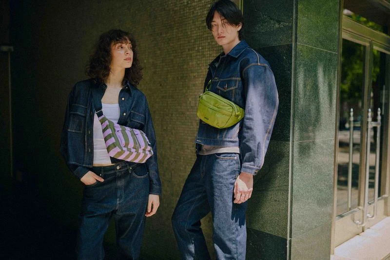 Two young adults, a man and woman, pose outdoors in matching denim outfits, each wearing a different color crossbody bag.