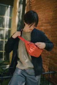 Man wearing a black bomber jacket over an olive sweater and grey cargo trousers holding a bright red fanny pack against a brick wall.