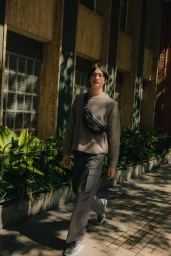 Model wearing a knit sweater and dark trousers carrying a black crossbody bag walking on a sunny city sidewalk past a building exterior