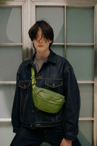 Young man in dark denim jacket wearing a bright green fanny pack against a gridded glass background