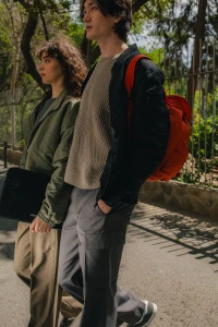 Two models walking outdoors wearing casual layered jackets and carrying a red backpack and black accessory case.