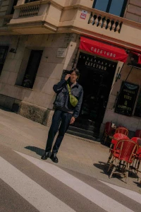 Man wearing a dark denim jacket and pants with a green crossbody bag standing on a street outside a cafe