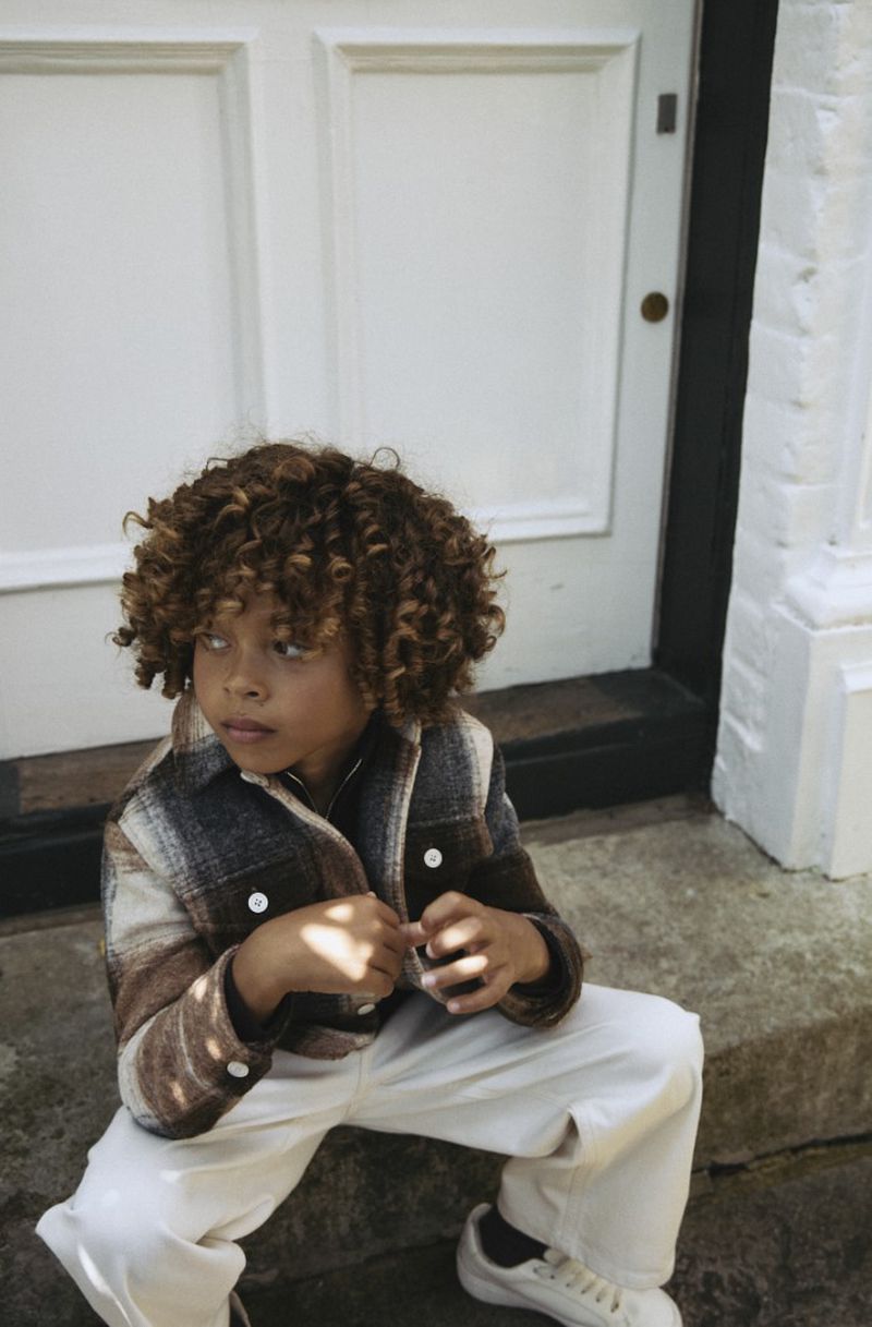Young child with curly brown hair sits on steps wearing a brown and white plaid jacket, cream-colored pants, and white sneakers in natural light.