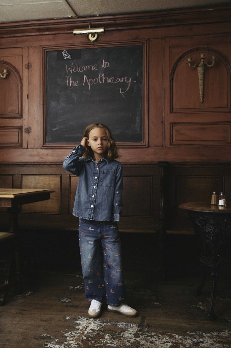 A girl in a denim shirt and patterned pants stands in front of a wooden wall with a chalkboard in an indoor setting.
