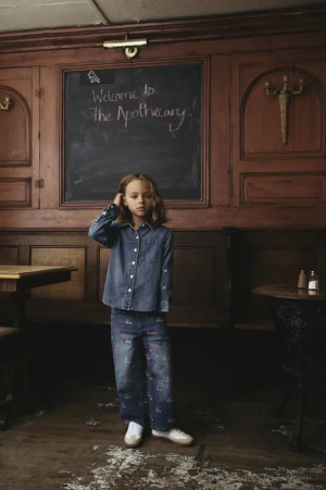 A girl in a denim shirt and patterned pants stands in front of a wooden wall with a chalkboard in an indoor setting.