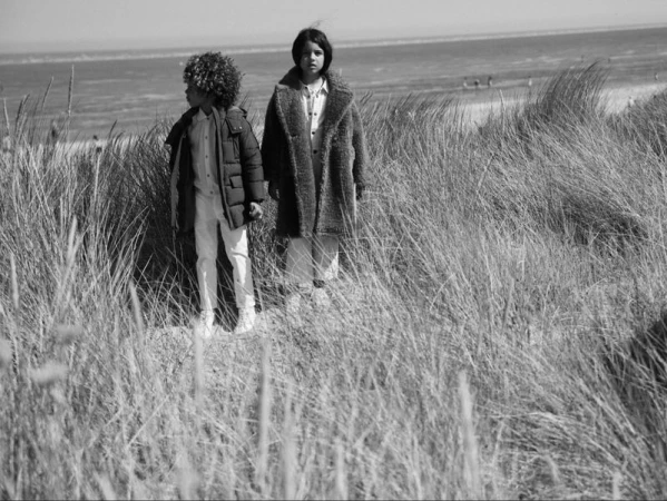 Two children in winter coats standing among tall beach grass near the ocean, photographed in monochrome.