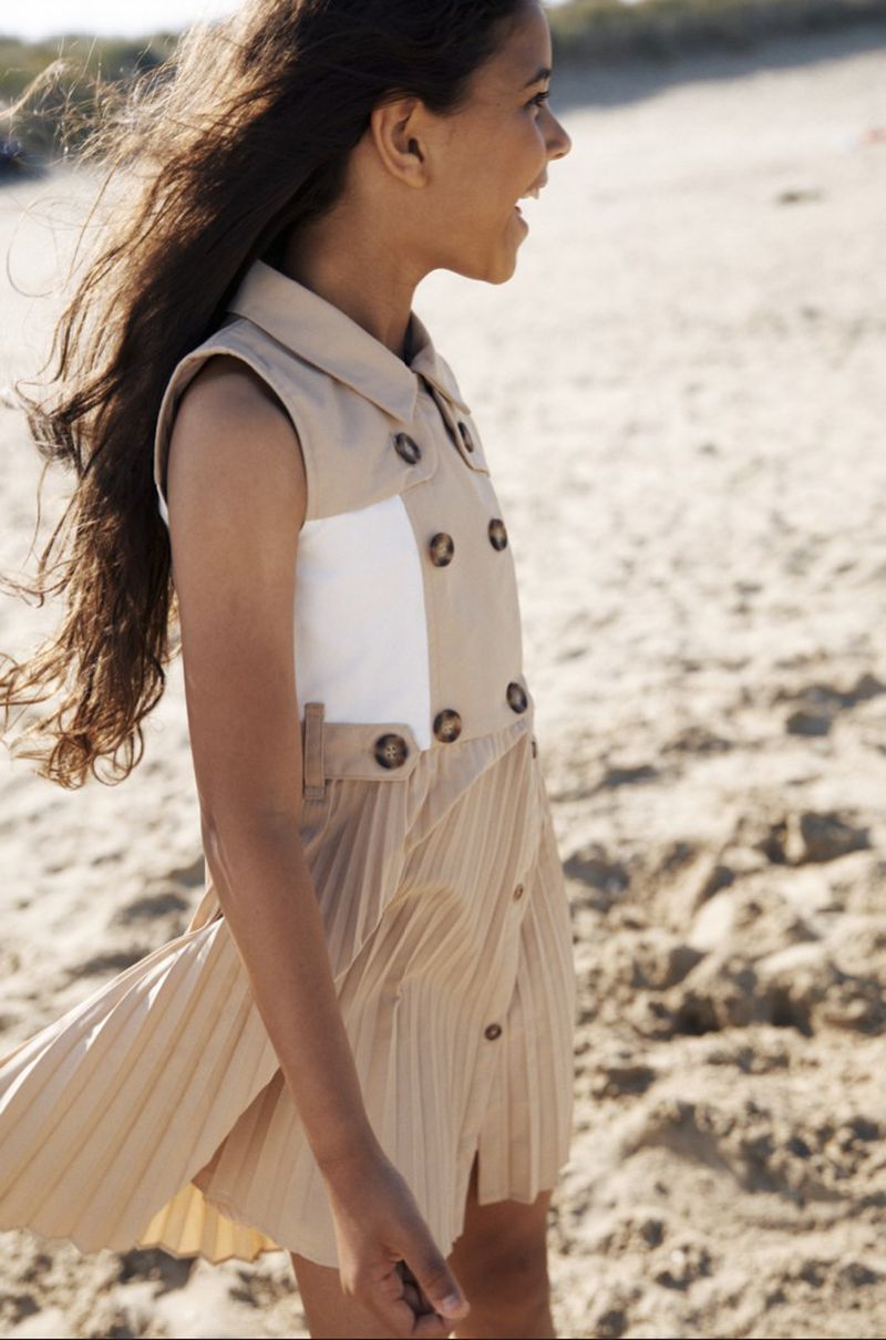 A young girl smiles while walking on a beach, wearing a stylish sleeveless dress with a pleated skirt and button detailing.