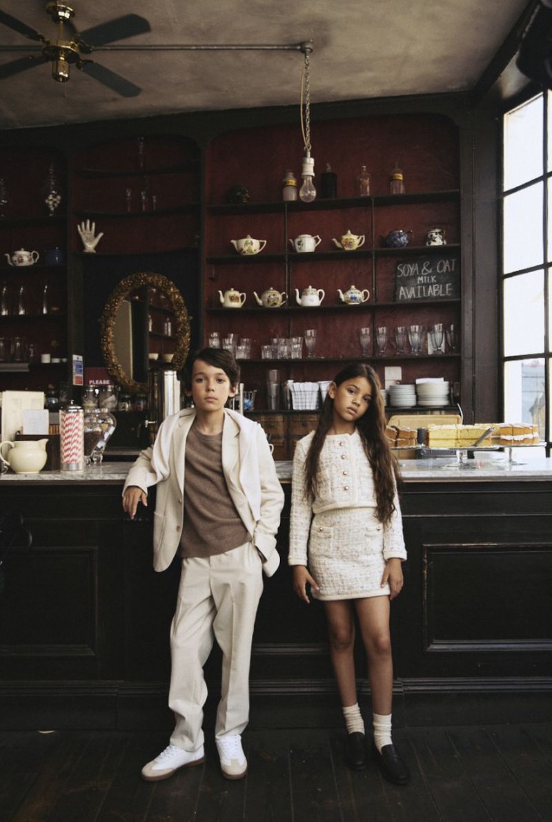 Stylish kids pose in vintage café: Boy in beige suit, girl in tweed skirt set, classic setting, high-fashion children's wear.