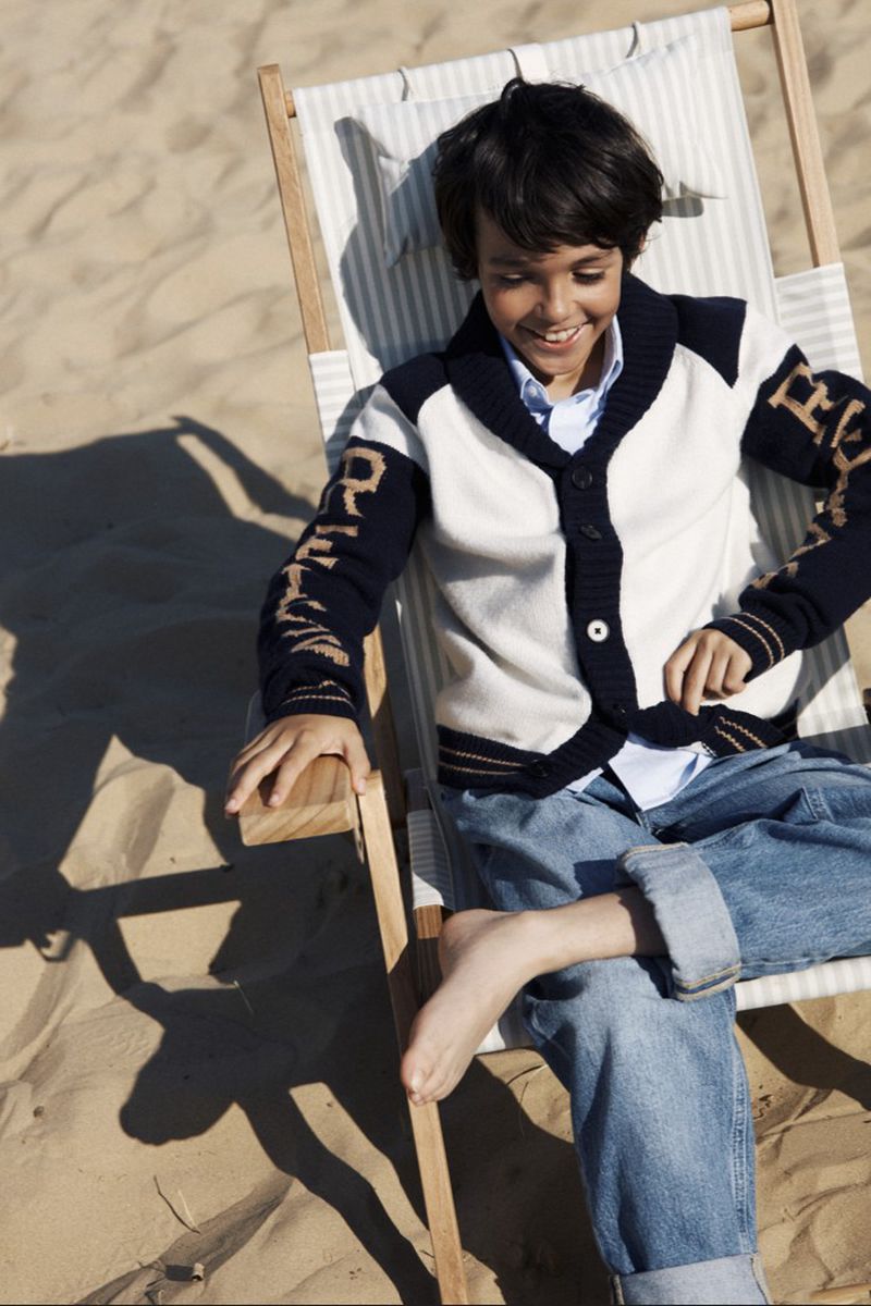 Boy in cardigan and jeans seated in a striped deck chair on sand, smiling in the sunlight, with a shadow on the beach.