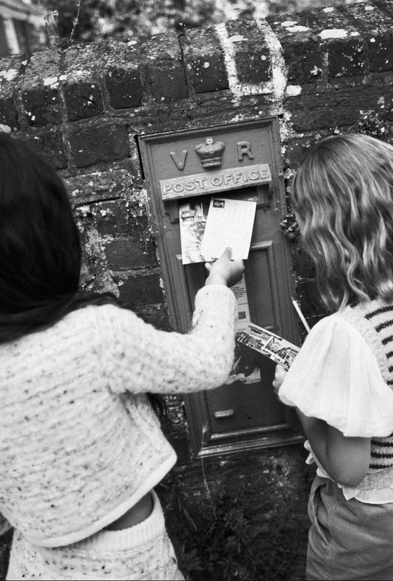 Two young girls mailing letters at a vintage-style UK post box set in a weathered brick wall, monochrome photo with shallow depth of field.