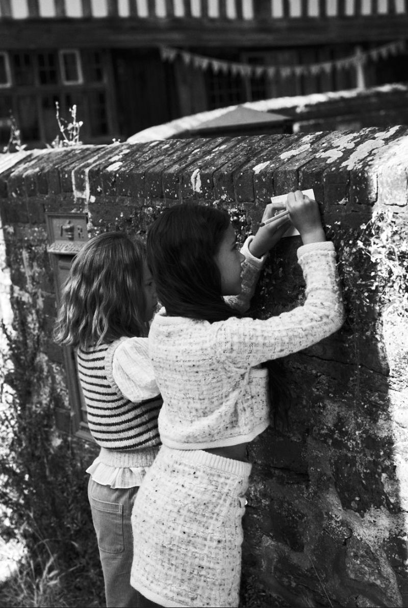 Two young girls interact with a brick wall, possibly posting or examining something, in an outdoor setting with a house in the background.