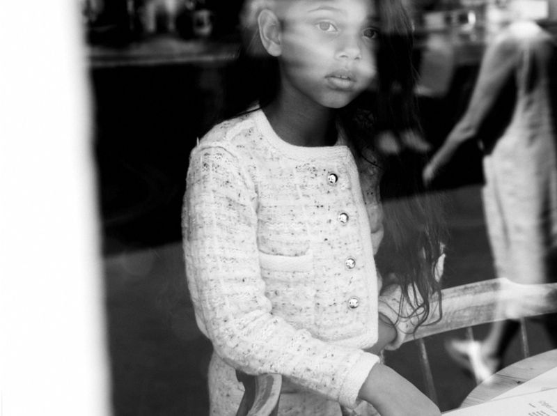 Black and white portrait of a young girl with dark hair and a white patterned cardigan, seen through a window reflection of a street scene.