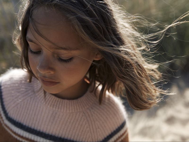Close-up of a young girl with brown hair wearing a striped sweater, looking down in a natural outdoor setting with soft, diffused lighting.