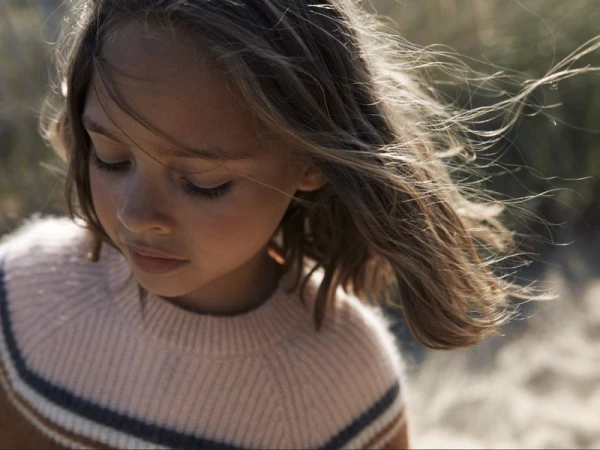 Close-up of a young girl with brown hair wearing a striped sweater, looking down in a natural outdoor setting with soft, diffused lighting.