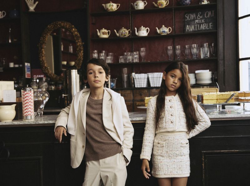A young boy and girl in matching outfits pose inside a restaurant with teapots and drink menu in the background, indoor shot.