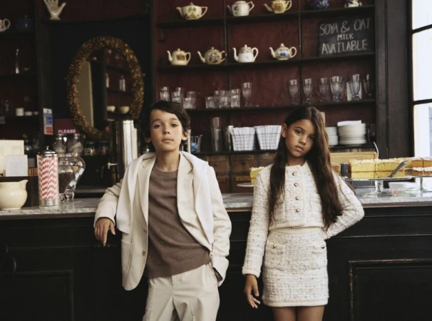 A young boy and girl in matching outfits pose inside a restaurant with teapots and drink menu in the background, indoor shot.