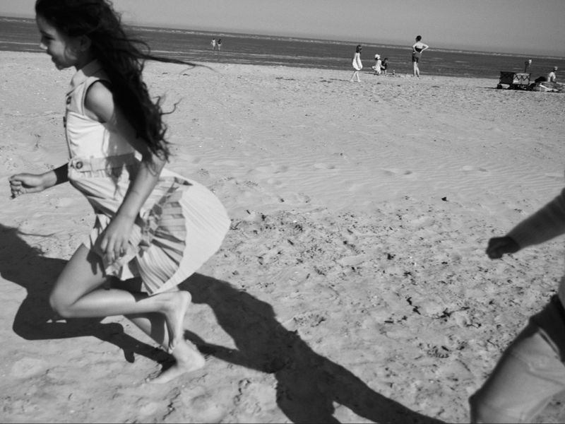 A young girl running barefoot on a sandy beach in a pleated dress with distant figures near the water in black and white.