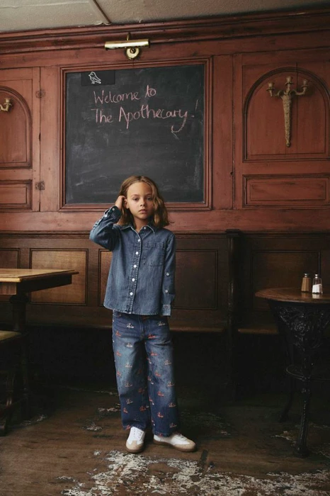 A child in a denim shirt and patterned jeans standing in a wood-paneled room with a chalkboard sign.