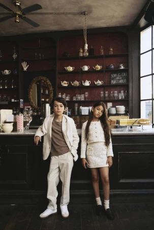 Boy in beige suit and girl in tweed skirt set posing inside a cafe with dark wooden shelves and red walls for Reiss Kids Fall 2024.