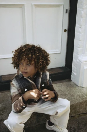 Young child with curly hair wearing a plaid jacket and white trousers sitting on stone steps outside a white door.