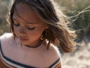 Close-up portrait of a child with hair blowing in the wind wearing a striped knit sweater outdoors.