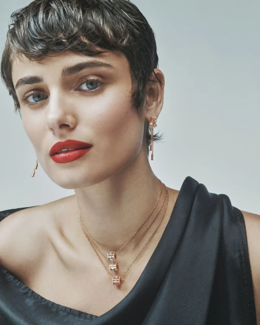 Close-up shot of an adult female with short brown hair, red lipstick, and blue eyes, wearing jewelry on a light-gray background.