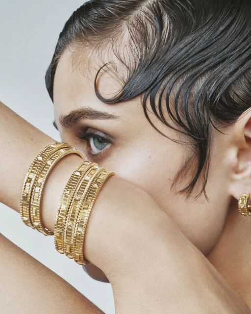 Close-up of a woman with sleek wet-look hair wearing golden, geometric-patterned bangle bracelets and a matching earring.