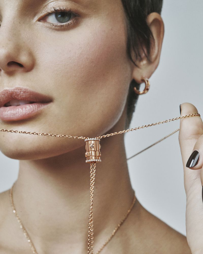 Close-up of a woman's face and gold necklace with diamond accents. The model has short dark hair, gold earrings, and neutral makeup.